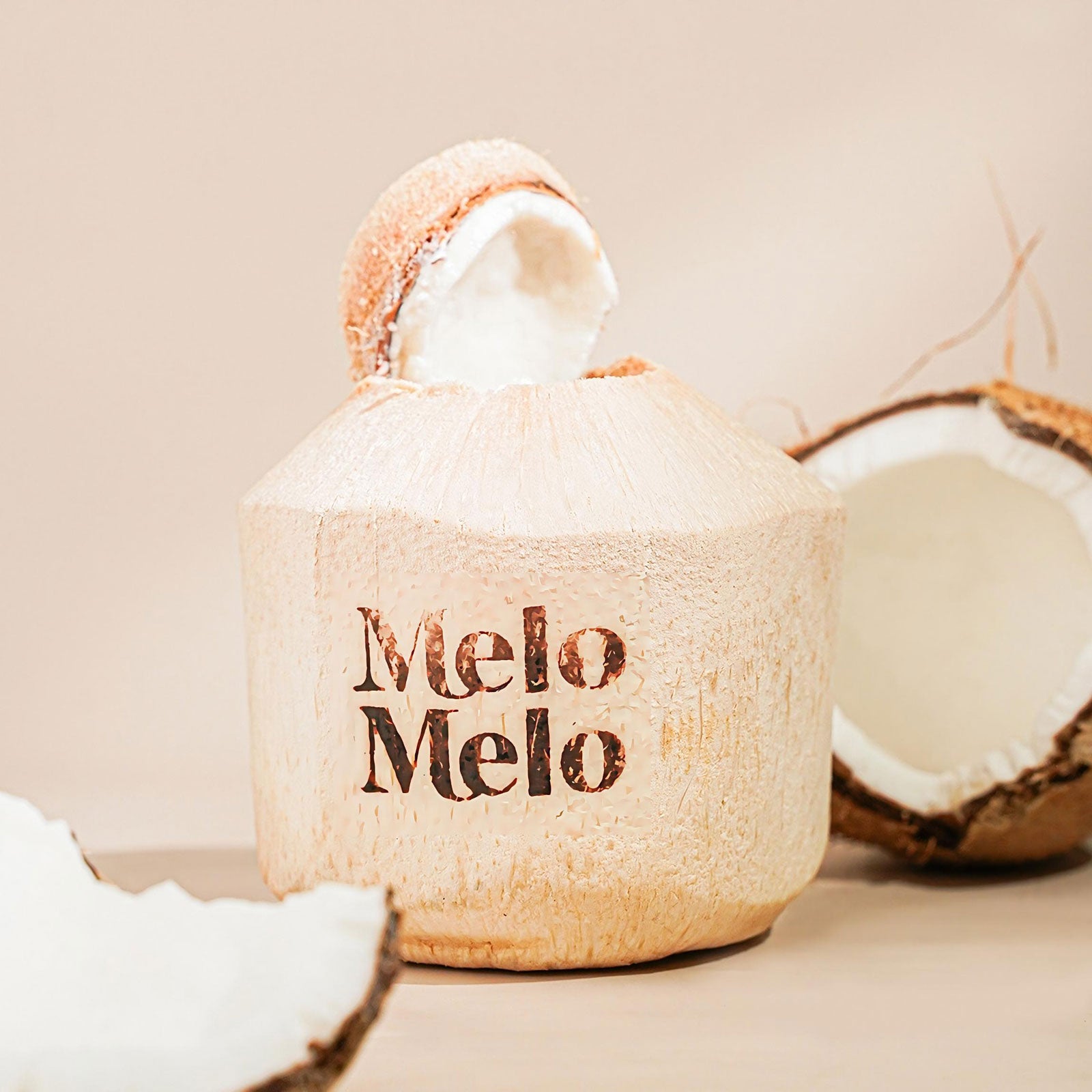 A fresh coconut with the top opened, revealing soft coconut flesh inside, branded with “Melo Melo” on the shell. Surrounded by natural coconut halves, the image features a clean, minimalist background with soft warm lighting, highlighting a tropical, premium, and refreshing aesthetic.
