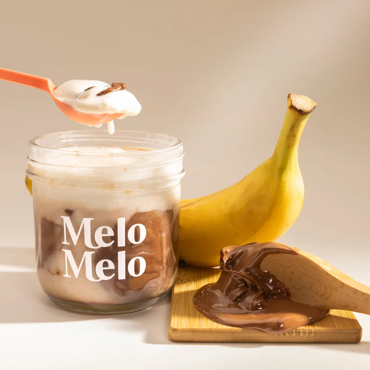 Banana Nutella Coconut coconut jelly in glass jar, creamy coconut layer with fresh banana slices and Nutella® hazelnut cocoa spread  base, spoon lifting silky jelly, dripping texture, fresh banana and Nutella® hazelnut beside, clean beige background, soft natural light, premium food photography